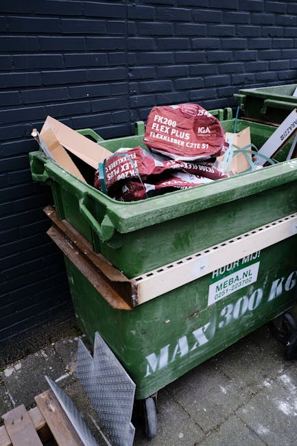 A waste collection worker dressed in a high-visibility yellow and red jacket is actively engaged in emptying a black garbage bag into the rear compactor of a large red rubbish truck, which is parked o