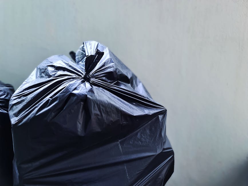 Several large black plastic garbage bags are stacked on the pavement near the edge of a street, with some appearing partially torn or crumpled. The bags contain domestic waste and are tied at the top,