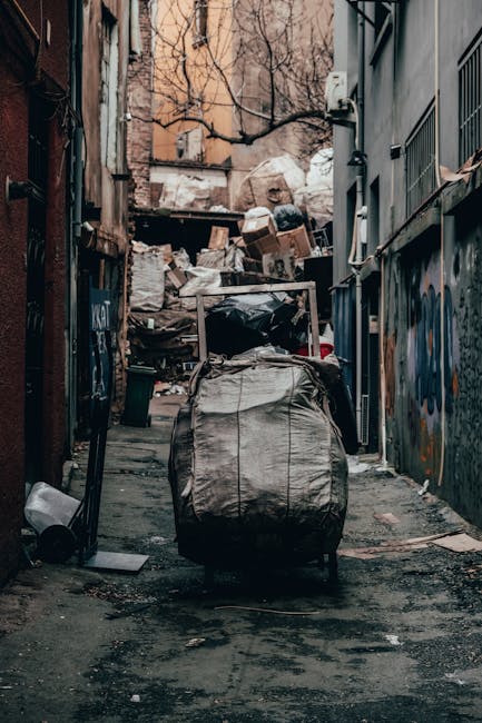 A large pile of mixed household waste and recyclable materials, including cardboard boxes, paper bags, plastic packaging, and black garbage bags, is gathered around three different color-coded rubbish