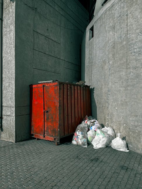 A close-up view of an overflowing pile of domestic rubbish accumulated on a paved area, including black plastic bin bags, a yellow plastic container, and a discarded car seat with fabric upholstery sh